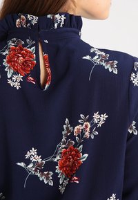 Námořnická květinová blůza se skládacím výstřihem, s červenými květy a bílými akcenty. Otvor na zádech ve tvaru klíče, měkká texturovaná látka.