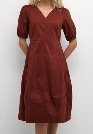 Kobieta w miętowej sukience midi w kolorze rdzy z krótkimi bufkami i dekoltem w serek, stoi z rękami za plecami.