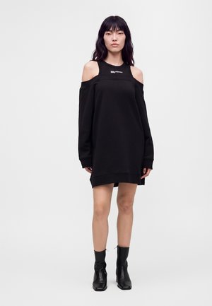 Giovane donna che indossa un vestito nero con le spalle scoperte e maniche lunghe e stivaletti neri alla caviglia, in piedi davanti a uno sfondo semplice.