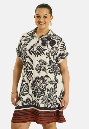 Femme souriante portant une robe courte à manches courtes longueur genou avec un motif floral noir et un ourlet rayé rouge, accessoirisée avec des boucles d’oreilles et un bracelet.