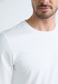 Tricou alb din bumbac cu guler rotund, textură netedă, design ajustat, cusături vizibile în jurul gulerului, fără modele sau grafică.