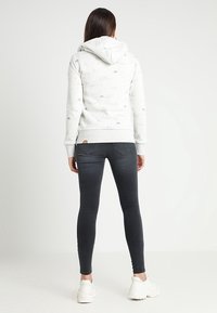 Vaalea harmaa huppari, jossa on käännettävät hihansuut, hienovarainen eläinkuvio ja pieni logopatchi, yhdistettynä tummiin kapeisiin farkkuihin ja valkoisiin lenkkareihin.