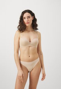 Nude bh med mjukt, slätt tyg, djup halsringning, justerbara band och delikat spetsdetalj; matchande bikinitrosor med hög skärning i liknande nyans.