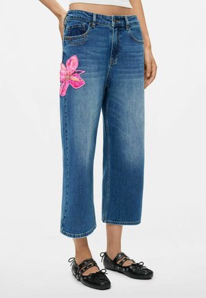 Blå denim cropped vidbenede jeans med en stor lyserød blomst patches på venstre lår, båret med sorte snørebånd flade sko med metal øjer.