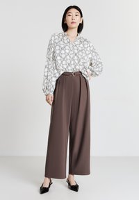 Donna in piedi con ampi pantaloni marroni e una camicia bianca con motivi geometrici a catena neri, indossando tacchi a punta nera.