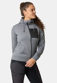 Kvinde med krøllet hår iført en grå lynlåshættetrøje med en sort brystlomme og sorte bukser, der kigger mod højre.