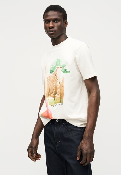 Noor mees, kes kannab valget T-särki, millel on kujutatud Vana-Egiptuse tempel, ja tumelisi siniseid teksaseid, seisab ühevärvilise valge tausta ees.