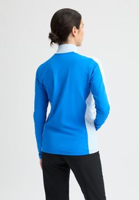 Camiseta deportiva de manga larga en azul brillante con paneles laterales blancos, confeccionada en tela elástica; cuenta con un cuello alto y un diseño ajustado.