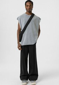 Grijze mouwloze shirt, zwarte losse broek en sneakers met kleuraccenten. Zwarte schoudertas. Effen achtergrond.
