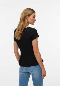 Sieviete ar gariem blondiem matiem, valkājot melnu īsām piedurknēm pieguļošu topu un zilas džinsas, stāv un skatās uz sāniem pret vienkāršu fonu.