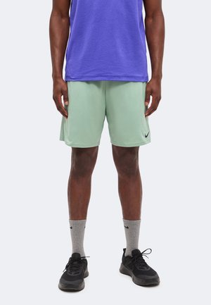 Hombre con pantalones cortos Nike de color verde claro, calcetines Nike grises, zapatillas negras y una camisa morada, de pie frente a un fondo claro y liso.