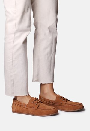 Brune ruskindsnørresko båret med cropped lyse beige bukser på en lys baggrund.