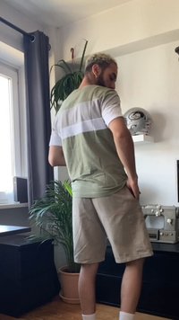 Kurzarmhemd mit olivgrünen und weißen horizontalen Streifen, kombiniert mit beigen Shorts. Inneneinstellung mit Zimmerpflanzen und einer Nähmaschine.