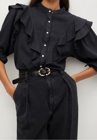 Blusa negra con detalles de volantes, cierre de botones y cuello alzado. Combinada con pantalones negros de cintura alta y un cinturón texturizado con hebilla dorada.
