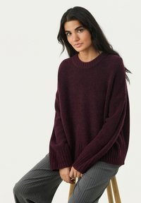 Maglione bordeaux, di taglia oversize, con collo e polsini a coste, realizzato in un tessuto a maglia testurizzato. Abbinato a pantaloni grigi a righe sottili.
