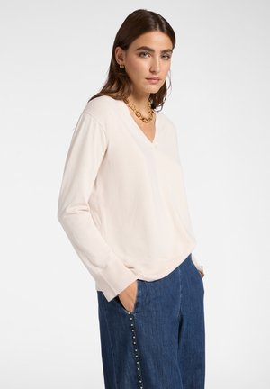 Donna in un maglione crema a scollo a V, jeans blu con borchie dorate, che indossa una collana d'oro e orecchini, mani nelle tasche, sfondo neutro.