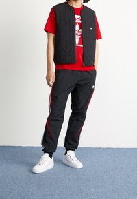 Svart vadderad väst över en röd t-shirt med grafik, ihop med svarta byxor med röda ränder och vita sneakers på en blå strukturerad yta.