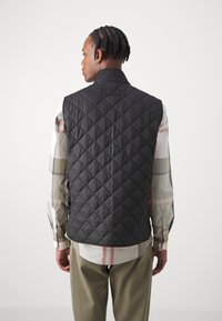 Colete preto acolchoado com gola alta, apresentando um aplique de logótipo nas costas. Usado sobre uma camisa xadrez multicolorida, combinado com calças verde-oliva claras.