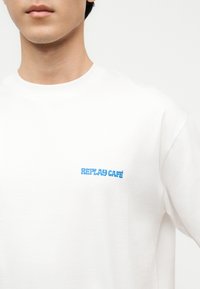 Νεαρό άτομο που φοράει λευκό φούτερ με μικρό μπλε κείμενο "REPLAY CAFÉ" στο αριστερό στήθος, μπροστά σε απλό λευκό φόντο.