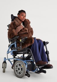 Fiatal nő barna bunda kabátban ül egy elektromos kerekesszékben, egy sima fehér háttér előtt.