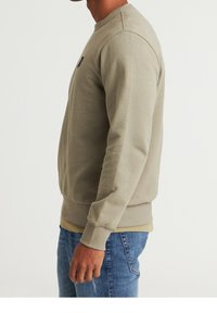 Sweatshirt beige à col rond en coton doux avec poignets et ourlet côtelés, présentant un petit logo noir sur la poitrine. Porté par-dessus une chemise de couleur claire.
