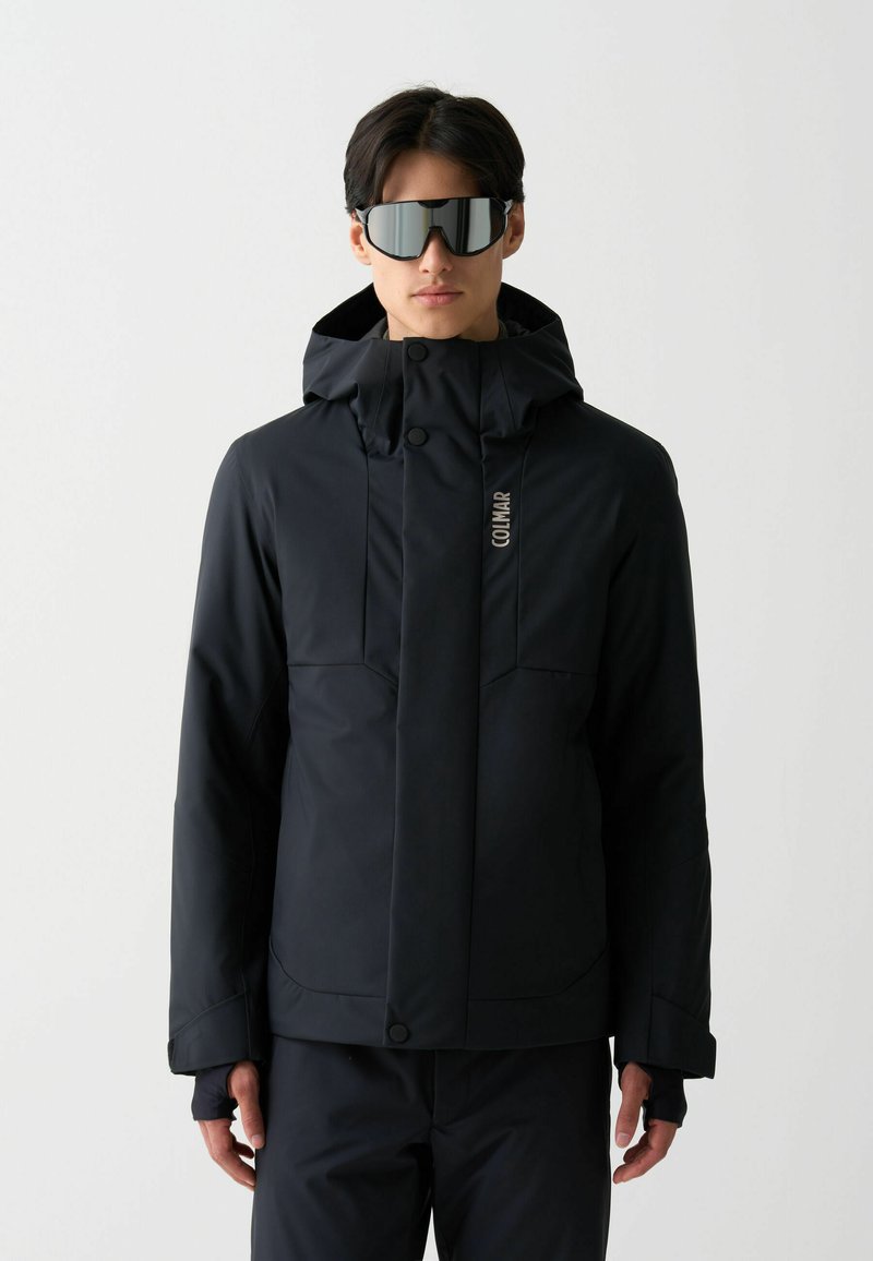 Svart vattentät jacka med huva och framdragkedjor, med en slät yta och en diskret logotyp på bröstet. Buren med reflexglasögon.