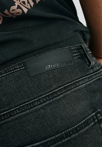 Czarne dżinsy z denimu z teksturowanym materiałem, widocznymi szwami oraz matową czarną skórzaną łatą z napisem "diverse" na pasku.