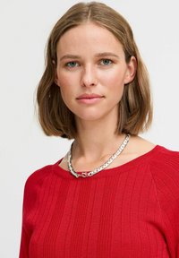 Silverfärgad kedjekedja med sammanlänkade länkar, bärs över en röd, ribbad långärmad topp med rund halsringning.