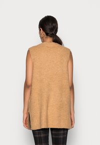 Beige ärmelloser Strickpullover mit gerippter Textur, seitlichen Schlitzen und Rundhalsausschnitt. Tragen über schwarzen karomusterhosen.