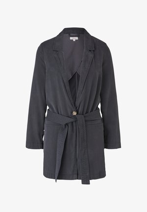 Kobieta ubrana w ciemną kurtkę z paskiem na wierzchu lawendowego prążkowanego topu i jasnoszarych dżinsów, stojąca z rękami swobodnie opuszczonymi wzdłuż ciała.