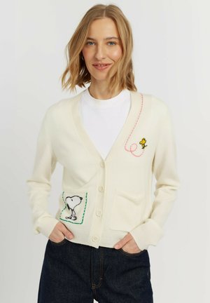 Blonde vrouw draagt een witte cardigan met Snoopy- en Woodstock-borduursel, gecombineerd met donkere jeans en een wit t-shirt.