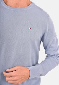 Maglione a maniche lunghe di colore blu chiaro, realizzato a maglia, con piccolo logo rettangolare rosso, bianco e blu marino sul petto, indossato da una persona con la mano vicino alla vita.
