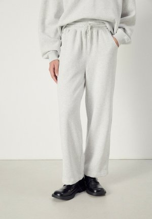 Persona che indossa pantaloni sportivi grigio chiaro con coulisse e scarpe di pelle nere, in piedi con una mano in tasca.