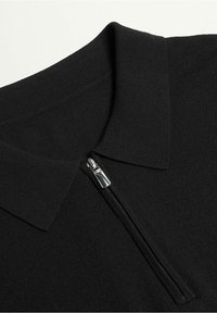 Polo noir avec col à fermeture éclair, fabriqué dans un tissu doux et texturé avec une tirette de fermeture éclair lisse. Vue rapprochée du col et de la fermeture éclair.