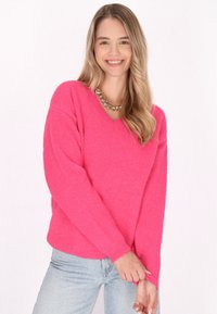 Pullover a maglia fucsia con scollo a V, maniche lunghe e polsini a costine. Indossato sopra jeans blu chiaro per un look casual.