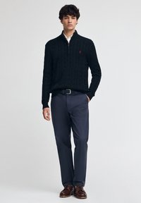 Pull noir en tricot torsadé avec une fermeture éclair quart et un logo brodé, associé à un pantalon bleu marine et des chaussures à lacets marron.