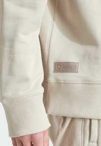 Béžový svetr s manžetami a lemem s žebrovaným úpletem a nášivkou loga týmu Mercedes-AMG Petronas Formula One na úrovni pasu, nošený s odpovídajícími kalhotami.