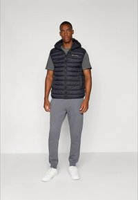 Muž v černé nafukovací vestě s kapucí, šedém tričku s krátkým rukávem, šedých teplácích a bílých teniskách stojí před jednobarevným pozadím.