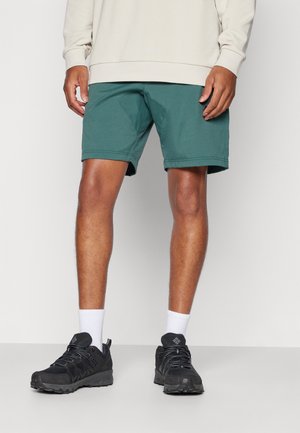 Calções atléticos verdes com uma textura suave, apresentando bolsos laterais e um corte confortável. Combinados com sapatilhas pretas e meias brancas até ao tornozelo.