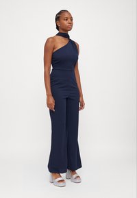 Macacão azul-marinho com design de um ombro, corpete justo e pernas largas, feito de um tecido suave. Combinado com sandálias de plataforma prateadas.