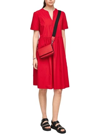 Femme portant une robe rouge longueur genoux à manches courtes, un sac à bandoulière rouge avec une sangle noire, des sandales noires et un bracelet clouté.