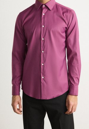 Homme portant une chemise ajustée à manches longues magenta avec des boutons blancs, associée à un pantalon noir, debout devant un fond uni.