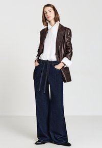 Blazer in pelle marrone scuro indossato su una camicia bianca a righe, abbinato a pantaloni in denim blu scuro a gamba larga con una cintura annodata. Texture morbide, design moderno.