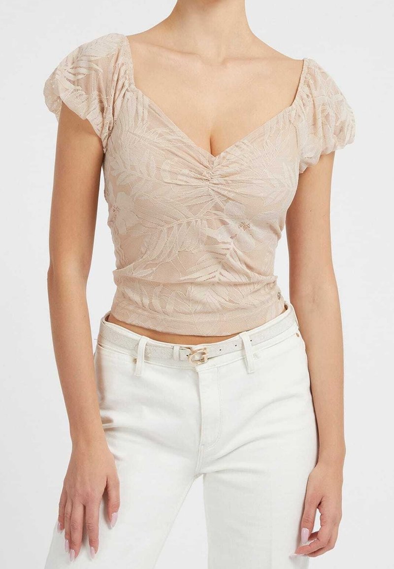 Femme portant un haut beige à manches courtes froncé en dentelle florale, associé à un pantalon blanc taille haute et une ceinture blanche.