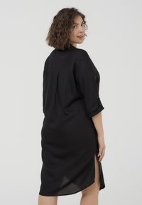 Abito nero con vestibilità comoda, maniche corte arrotolate, dettagli con piega sulla schiena e doppi spacchi laterali. Tessuto liscio, lunghezza sopra il ginocchio.