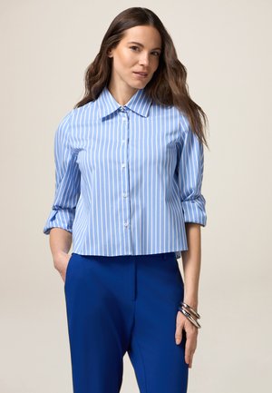 Camicia a maniche corte a righe blu chiaro con colletto e chiusura a pulsante, abbinata a pantaloni blu scuro e bracciali d'argento.