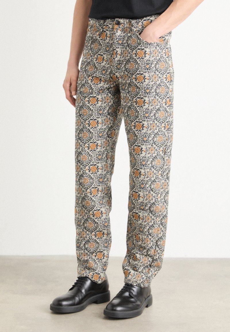 Personne portant un pantalon à motifs avec un design floral orange et gris, un haut noir et des chaussures en cuir noires à lacets. Une main dans la poche.