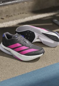 Ζεύγος μαύρα και ροζ παπούτσια Adidas για τρέξιμο σε υφή εξωτερικής επιφάνειας κοντά σε μπλε γραμμή.