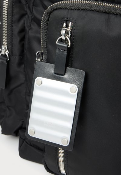 Sac à dos en tissu noir avec des fermetures éclair argentées et une étiquette de bagage en cuir noir avec une plaque blanche vierge attachée.