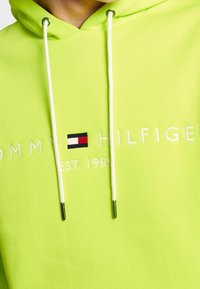 Felpa verde lime con cappuccio e lacci bianchi, con un logo ricamato in rosso, bianco e blu. Il testo recita "TOMMY HILFIGER EST. 1985."